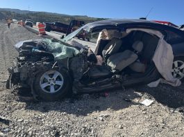 Dark SUV with severe front-end damage and deployed airbags on a gravel shoulder, traffic cones and responders in the distance on a sunny highway construction scene.