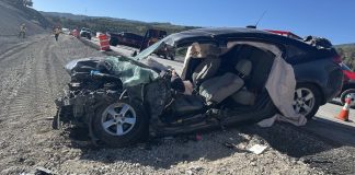 Dark SUV with severe front-end damage and deployed airbags on a gravel shoulder, traffic cones and responders in the distance on a sunny highway construction scene.