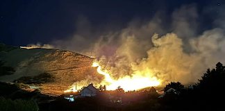 Nighttime wildfire on a hillside with bright flames, smoke rising above a small residential area.