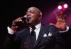 Peabo Bryson, singer known for Disney hits, dies at 75 after stroke Male singer in a dark suit performs on stage, microphone in hand, pink spotlight behind him.