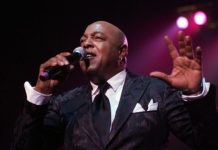 Male singer in a dark suit performs on stage, microphone in hand, pink spotlight behind him.