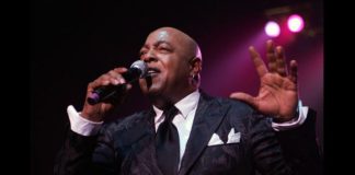 Male singer in a dark suit performs on stage, microphone in hand, pink spotlight behind him.