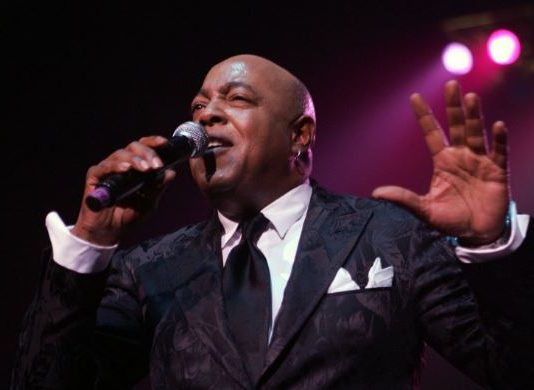 Male singer in a dark suit performs on stage, microphone in hand, pink spotlight behind him.