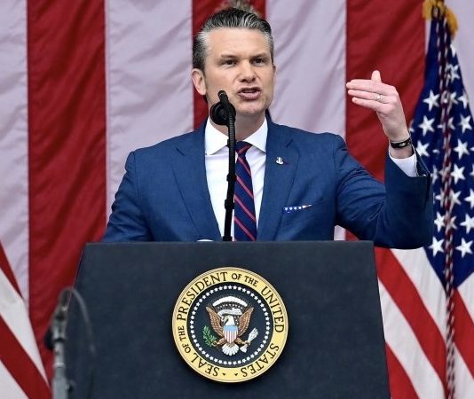Man in a blue suit speaking at a podium with the presidential seal, flanked by U.S. flags behind him.