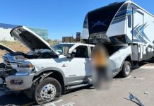 White RAM pickup with its hood open beside a large fifth-wheel RV in a parking lot; a blurred person stands between them.