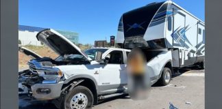 White RAM pickup with its hood open beside a large fifth-wheel RV in a parking lot; a blurred person stands between them.