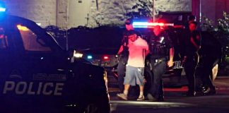 Night scene with police car and officers detaining a man beside flashing red and blue lights.