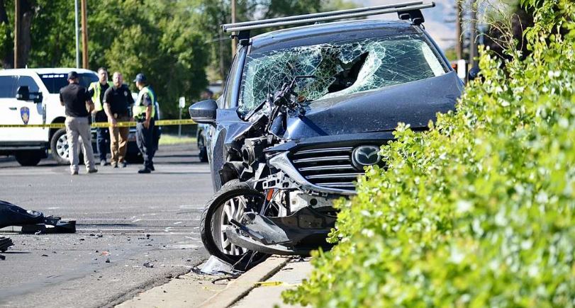 Blue SUV with severe front-end damage at curb after crash; police and responders in background, scene cordoned off with yellow tape.