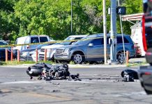 Motorcycle wreck with scattered debris on a city street, behind yellow caution tape and parked cars in background.