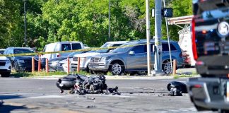 Motorcycle wreck with scattered debris on a city street, behind yellow caution tape and parked cars in background.