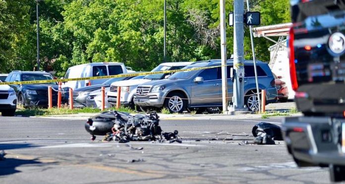 Motorcycle wreck with scattered debris on a city street, behind yellow caution tape and parked cars in background.
