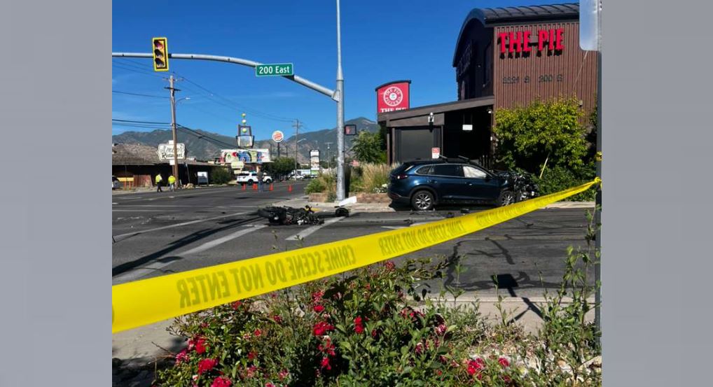 Accident scene at a street corner with yellow crime-scene tape, a crashed motorcycle, and a car near a restaurant named 'The Pie' on a sunny day.