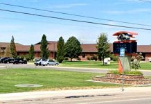 Wide view of a campus entrance: a brick building, parked cars, manicured lawns, and a prominent decorative sign on a stone island by the road under a blue sky with flags overhead.