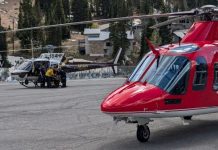 Red helicopter in the foreground on a paved tarmac, with a gray helicopter and ground crew nearby and a small alpine town with trees and buildings in the background.