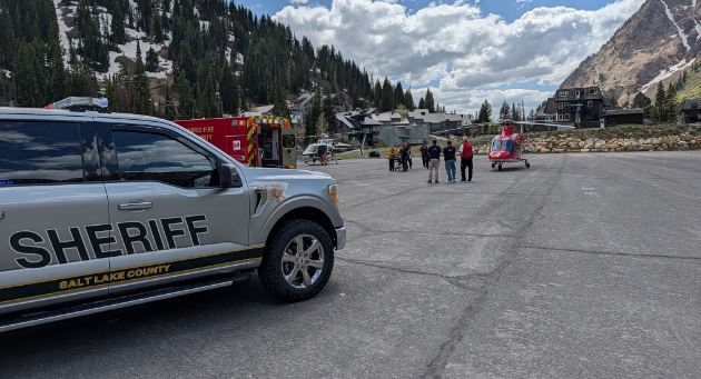 Sheriff's SUV in the foreground with a fire truck and a helicopter behind responders on a tarmac, mountains in the background.
