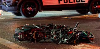 Motorcycle wreck on a street, laid on its side with debris; a police vehicle looms over the scene with flashing lights in the background.