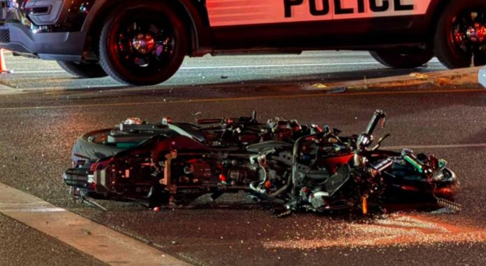 Motorcycle wreck on a street, laid on its side with debris; a police vehicle looms over the scene with flashing lights in the background.