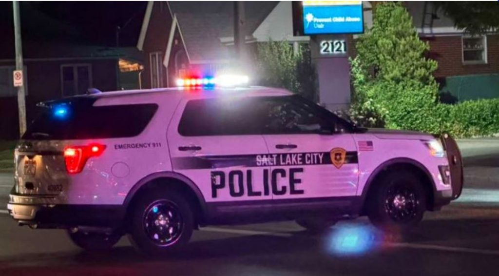 Salt Lake City Police SUV with red and blue emergency lights flashing at night parked on a street with a building and greenery in the background