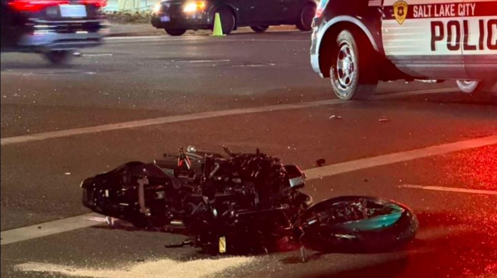 Wrecked motorcycle on a city street at night with debris around police car with Salt Lake City markings and red emergency lights in the background