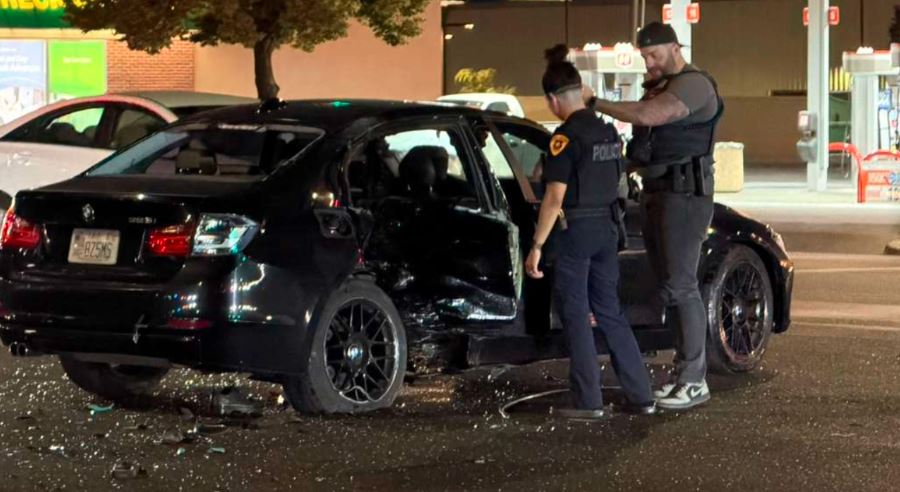 Two police officers inspect a heavily damaged black sedan with shattered glass on a nighttime street outside a store Note If needed shorten to a single sentence for screen readers