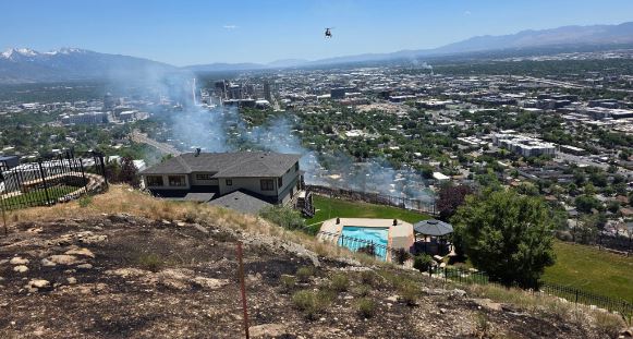 Hillside house with smoke rising from a brush fire a backyard pool and a helicopter overhead over a city valley
