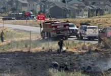 Firefighters spray water on a burned field near a neighborhood; a 'WRONG WAY' sign is visible by the road.