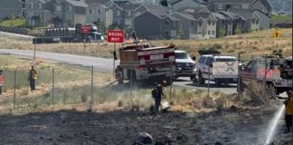 Firefighters spray water on a burned field near a neighborhood; a 'WRONG WAY' sign is visible by the road.