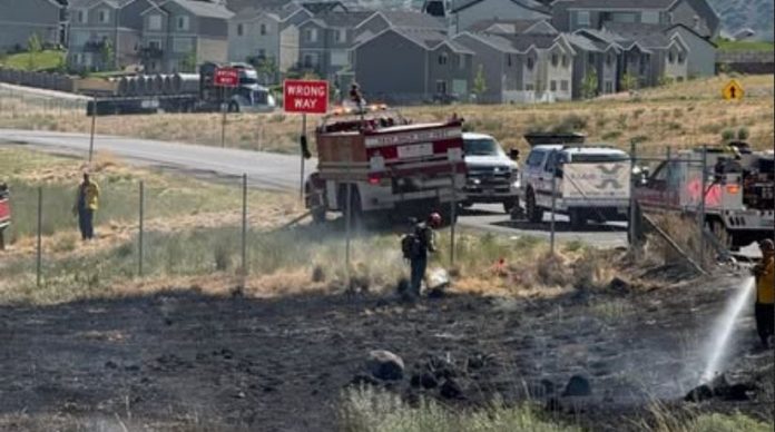 Firefighters spray water on a burned field near a neighborhood; a 'WRONG WAY' sign is visible by the road.