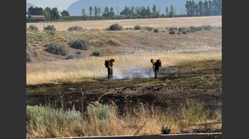 Two people wearing protective clothing tending a small brush fire in a dry field, with smoke rising and distant hills in the background.