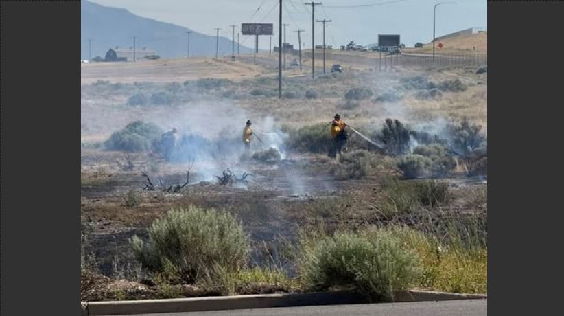 Two firefighters in yellow gear battle a brush fire in a dry desert area beside a road, smoke rising from the flames.