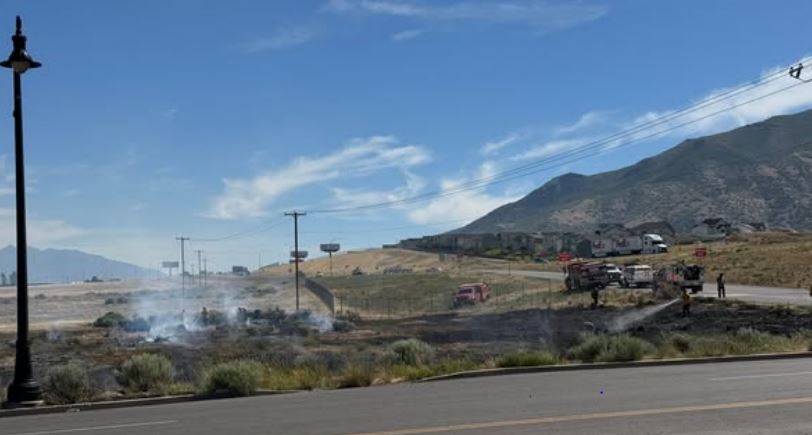 A roadside brush fire with smoke rising, emergency vehicles and responders on a two-lane road near a mountainous landscape.
