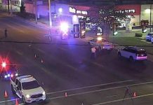 Nighttime intersection with police cars, flashing red and blue lights, and orange traffic cones blocking the road in front of a storefront.