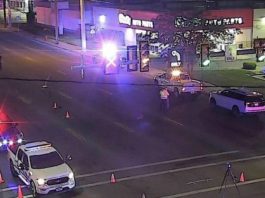 Nighttime intersection with police cars, flashing red and blue lights, and orange traffic cones blocking the road in front of a storefront.