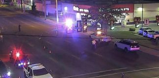 Nighttime intersection with police cars, flashing red and blue lights, and orange traffic cones blocking the road in front of a storefront.
