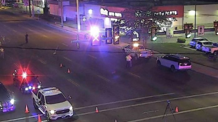 Motorcycle tow strap crash Nighttime intersection with police cars, flashing red and blue lights, and orange traffic cones blocking the road in front of a storefront.