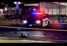 Police car with red and blue lights on at night behind yellow caution tape; a fallen bicycle lies on the wet road in the foreground.