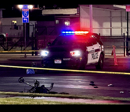 Police car with red and blue lights on at night behind yellow caution tape; a fallen bicycle lies on the wet road in the foreground.