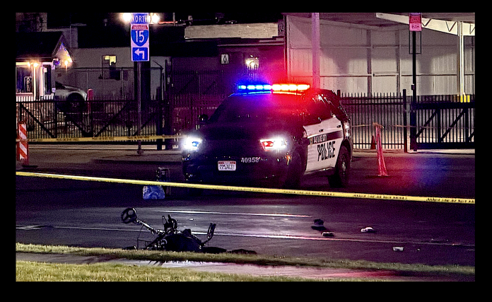 Police car with red and blue lights on at night behind yellow caution tape; a fallen bicycle lies on the wet road in the foreground.