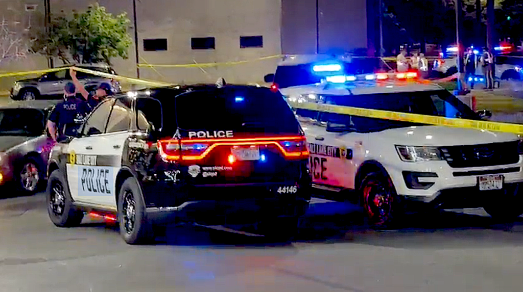 Nighttime police scene with two police SUVs, blue and red lights flashing, and yellow caution tape across a street at an incident site.