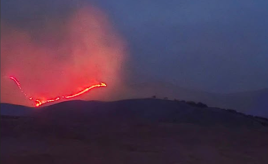 The South Mountain Fire in Tooele County | Gephardt Daily Lava flow down a hillside at dusk, with bright red molten rock glowing against a dark sky