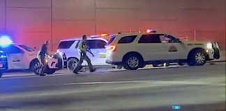Nighttime emergency scene with police car flashing blue lights, a white SUV, and responders near a pink-lit wall on a street.