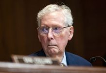 Sen. Mitch McConnell hospitalized, spokesman says Older man with gray hair and glasses in a dark suit sits at a desk in a formal hearing room, looking serious.