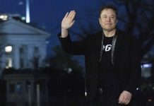 A man in a black jacket waving at night, standing outdoors near a lit neoclassical building.
