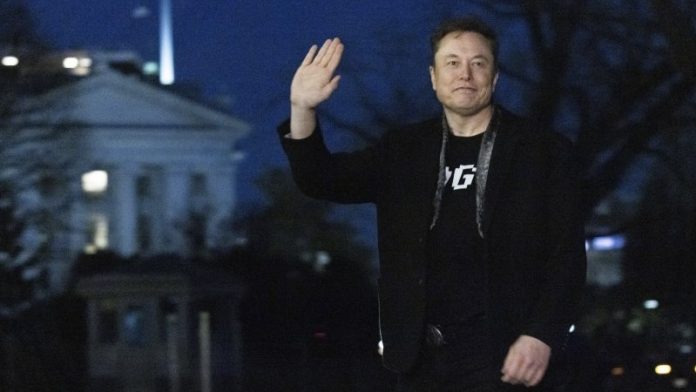 A man in a black jacket waving at night, standing outdoors near a lit neoclassical building.