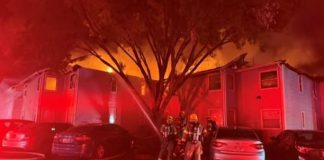 Firefighters in gear stand outside a smoke-filled, multi-unit building at night as flames glow red; cars are parked nearby and hoses cover the ground.