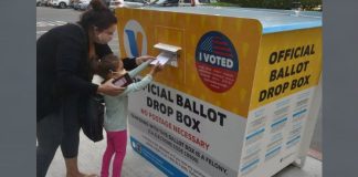 Mother and young girl deposit a ballot into a yellow official ballot drop box on a sidewalk, voter turnout visible on the box.