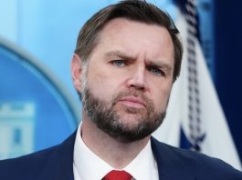 Bearded man in a dark suit and red tie with a serious expression against a blue backdrop.