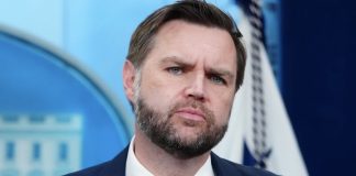 Bearded man in a dark suit and red tie with a serious expression against a blue backdrop.