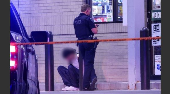 Police officer standing behind orange crime-scene tape while a kneeling person sits against a store wall outside a shop; scene appears to be an investigation area.