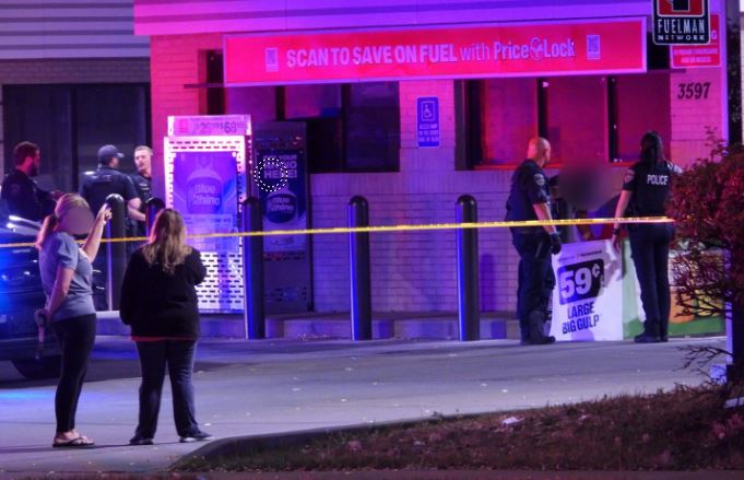 Police officers and bystanders stand behind yellow caution tape outside a gas station, with blue/purple lighting and a storefront visible in the background.
